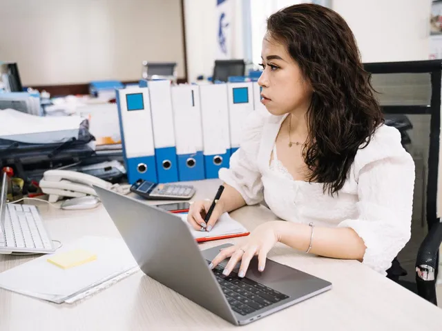 Woman working at a desk with a laptop, paperwork and binders.