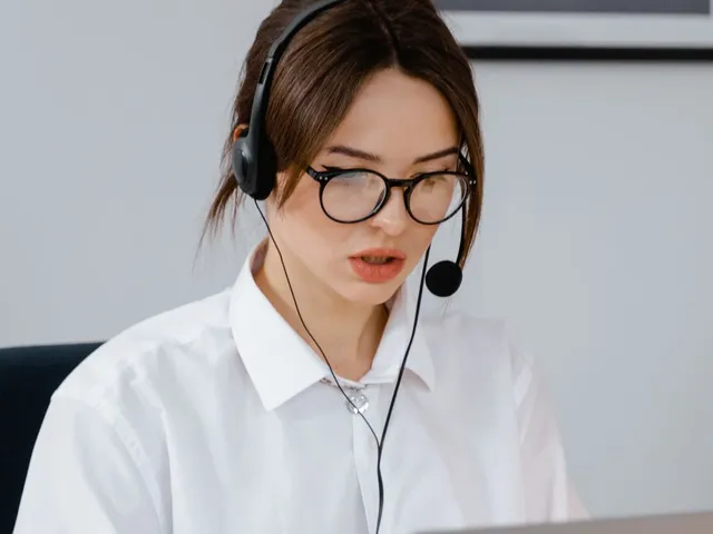 Woman in glasses talking on a headset while working on her laptop.