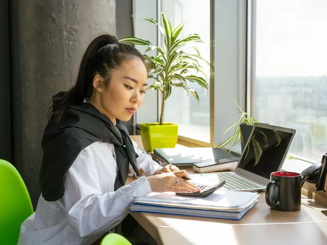 Woman working with a calculator, papers and a laptop by a window.